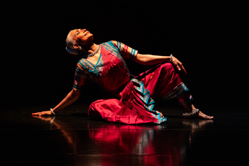 Bijayani Satpathy, seated, rests her weight on one buttuck and her arm, as she leans back but faces the light abover her. She is radiant, and composed, glowing in her bright red silk dress with details of blue and gold. 