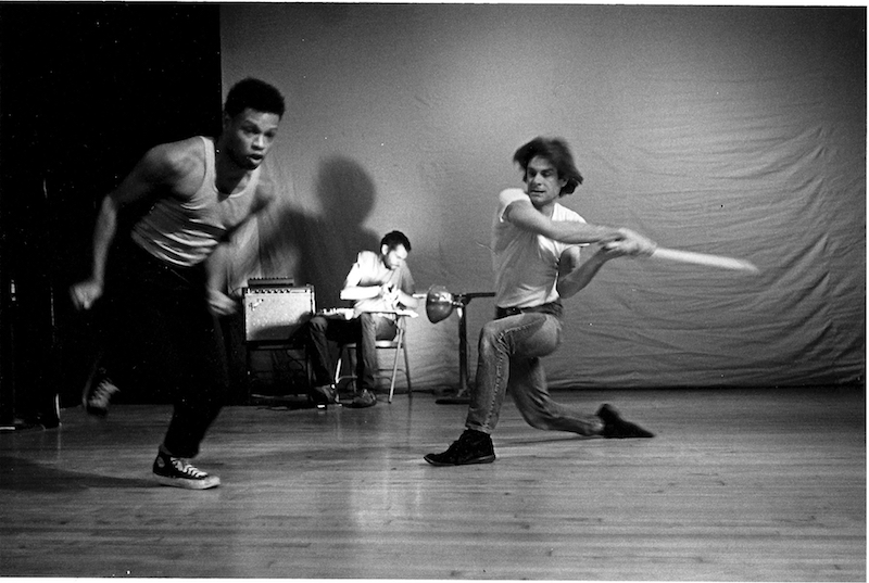 A woman in a tank top and sneakers in mid run while another performer kneels and swings a board. Someone sits with a guitar in the background.