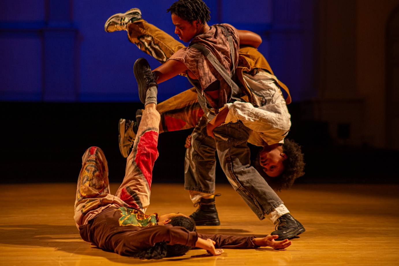 Three performers - one holding another upside down and the third lying on the floor under them with leg extended.
