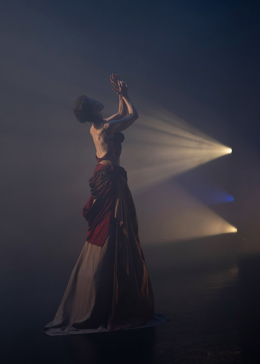 a beam of light radiates outward from a point at the side of the stage. It's light illuminates an otherwise darkly lit woman. We notice the musculature of her back and the flowing drapery of her skirt. 