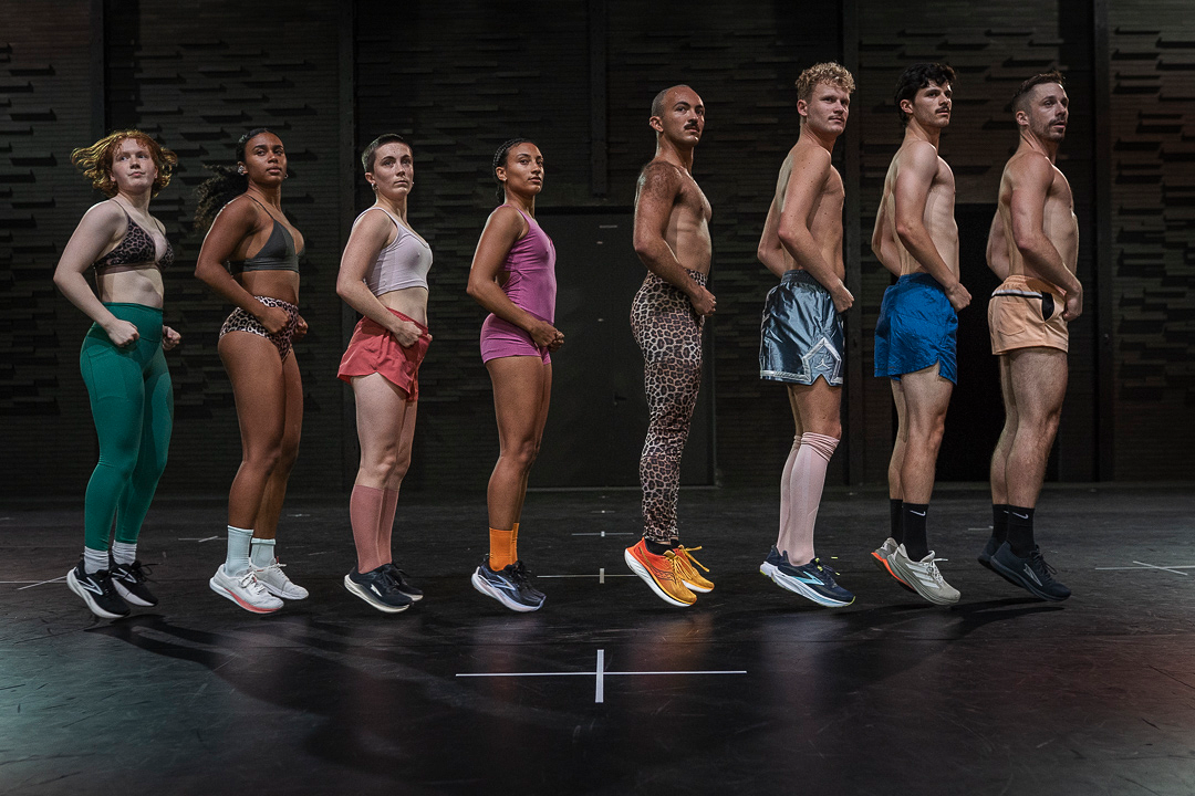 Eight dancers next to one another bodies in profile facing the audience  - four women of the same height followed by four men of the same height. Dressed in work out clothes, the dancers press their fists into their pelvis, as their heels and toes rise slightly off the floor..