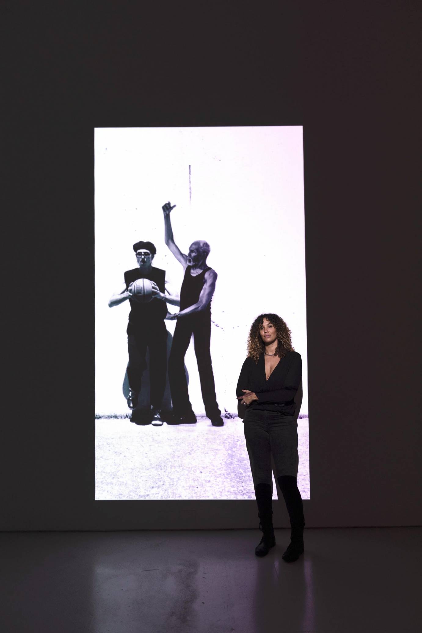 Curley Haired Choreographer Janessa Clark dressed totally in black stands in front of one of the black and white duets of her film communion.
