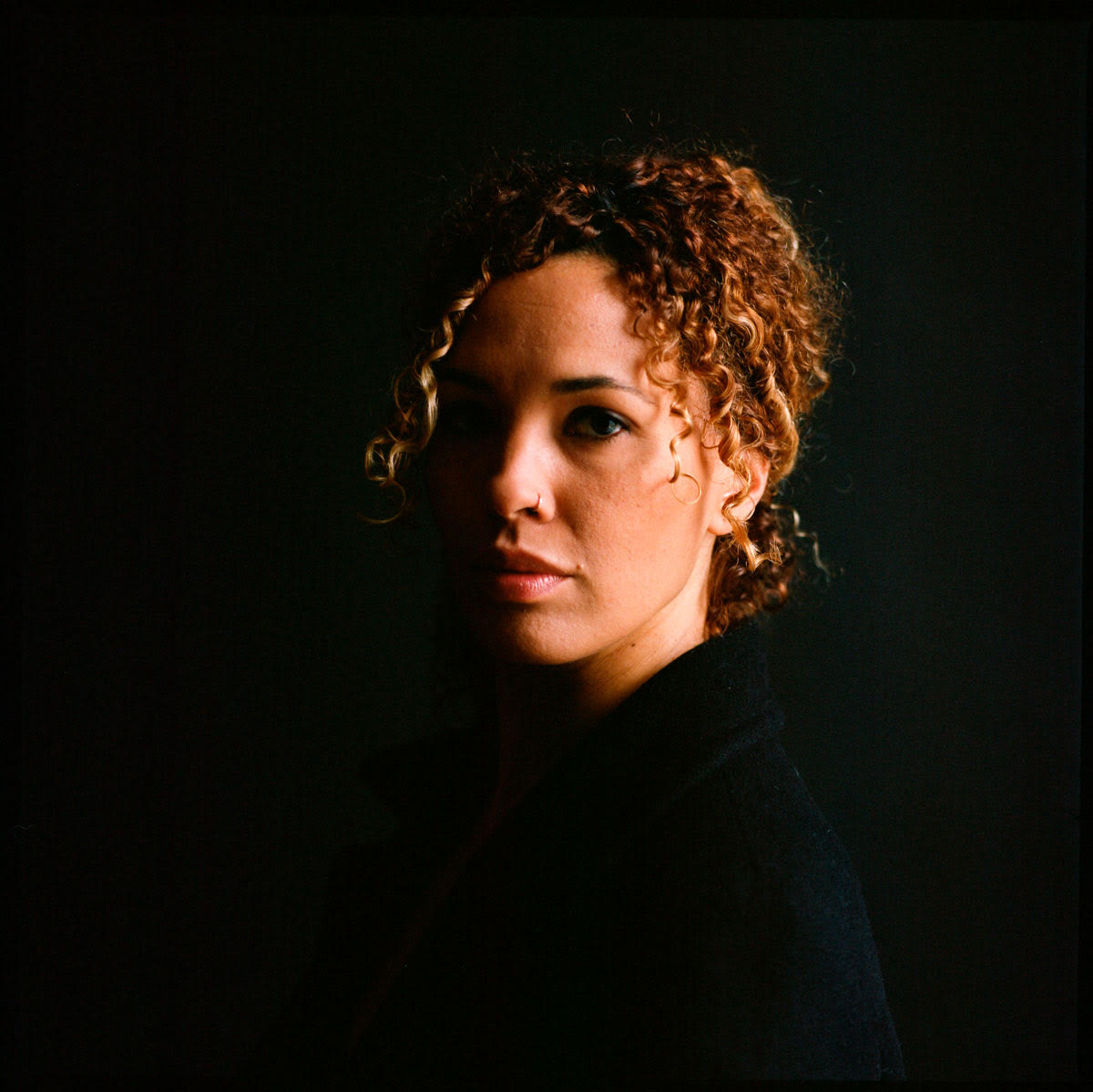 Janessa Clark, portrait, face partially in shadow,curly brown reddish hair with blonde accents tied back, she serenely looks out at us.
