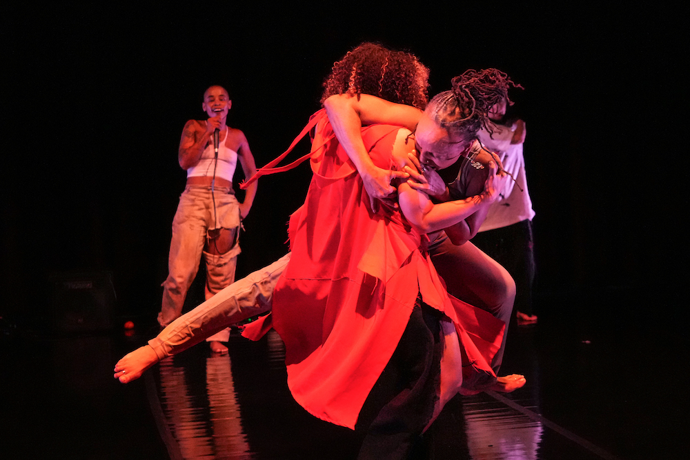 Charmaine Warren faces us and Dominica Greene in red has her back to us as the two embrace in an off balance pose, in the background Jasmine Hearn speaks or sings into a mic, as she observes the two dance