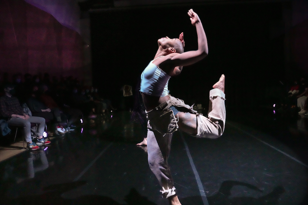 Jasmine Hearn in tight blue top midriff exposed,arms and head lifted up,wearing tattered light brown pants she stands in a balance with one leg bent behind her as the audience looks on