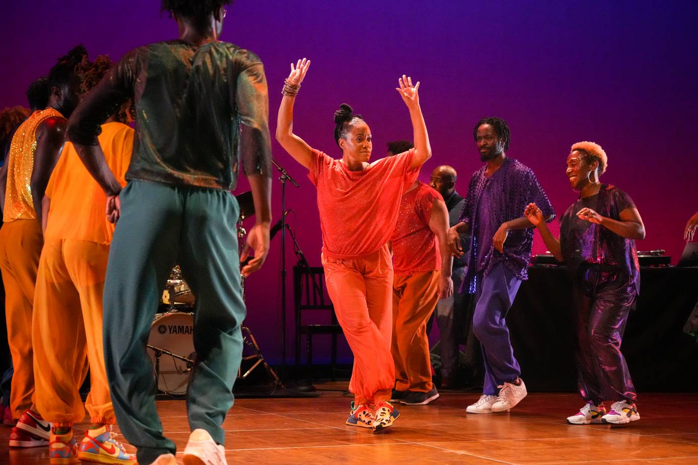 The group of Black Dancers in Rainbow colors form lines around a woman dancing in orange...