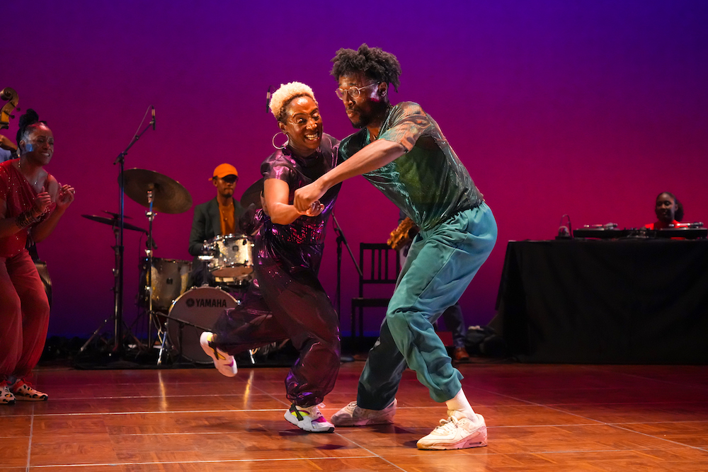 a Black couple dance together in front of band as another member of the group in orange cheers on