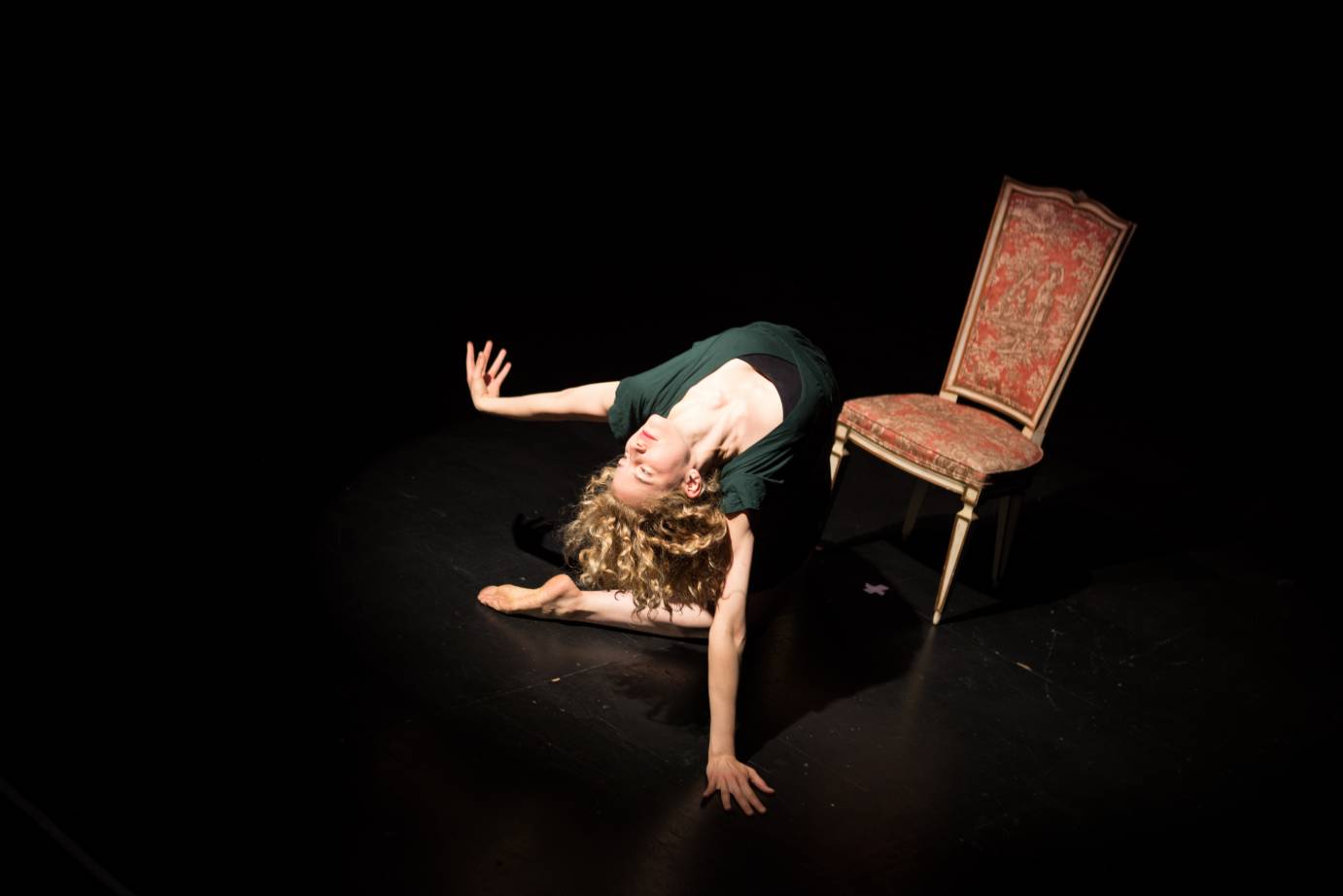 Beside an ornate chair, a woman sits on her knees in a deep backbend