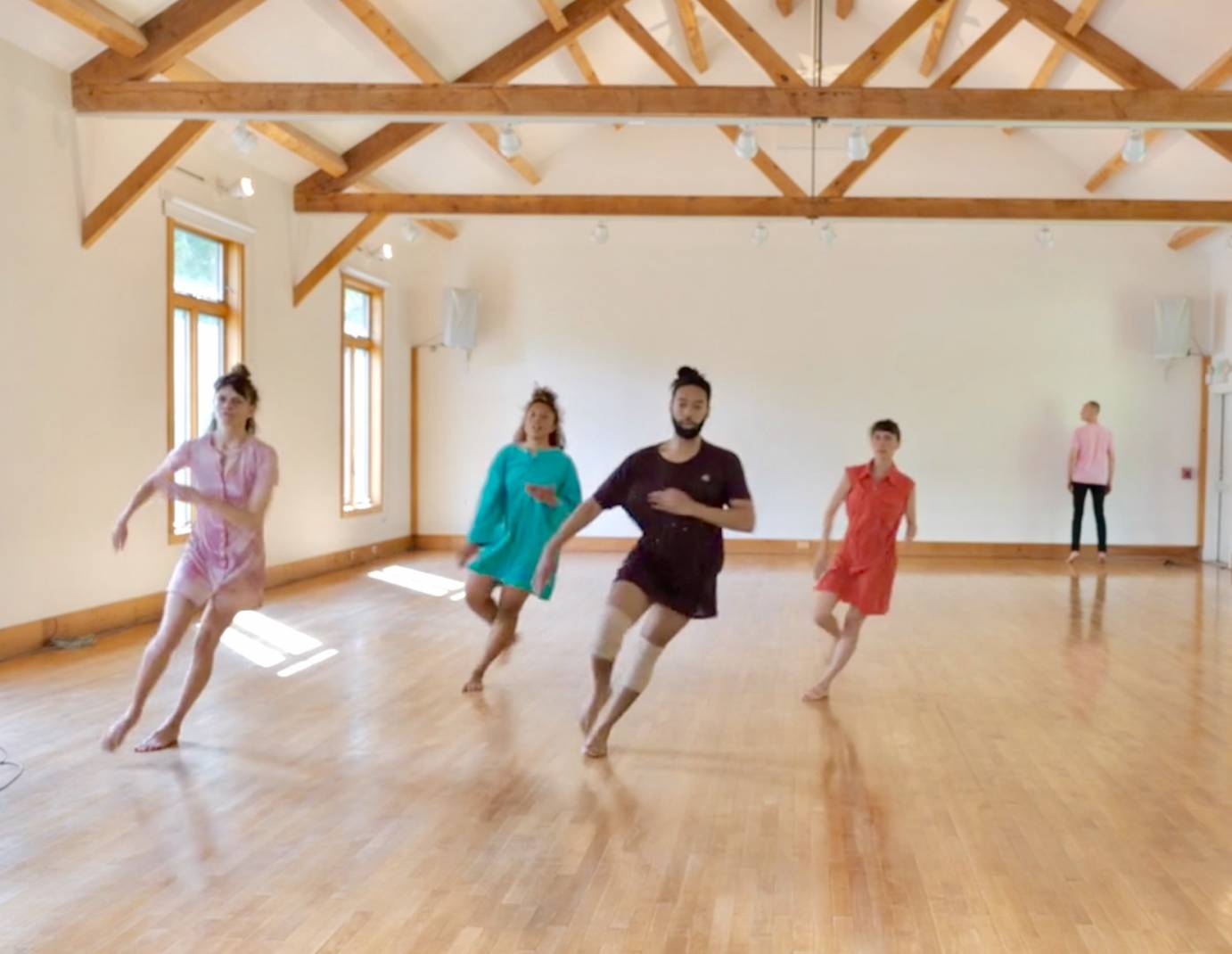 4 dancers wearing colorful and short outfits dance in a white-walled room with a light colored wood floor , one person in a pink top with black pants stands in the background 