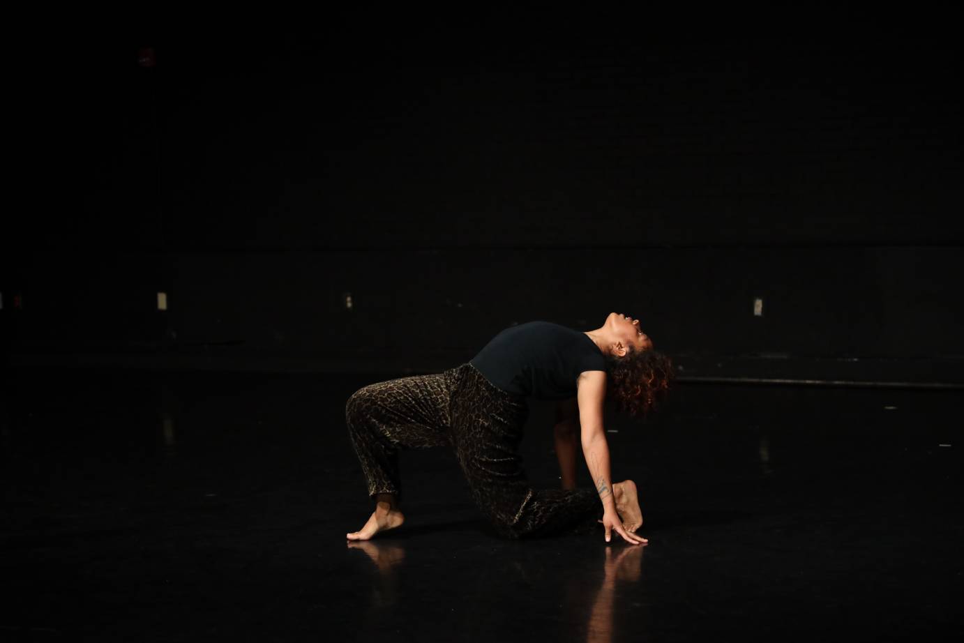  a brown-skinned woman in black space wearing a black t shirt and waht looks to be leopard print pants , arches her body while simultaneously mastering a kneeling lunge
