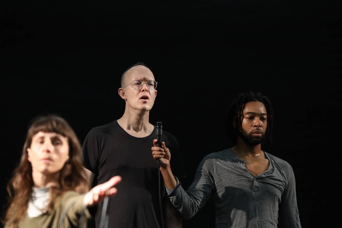 a trio consisting of  ( from l to r) an out of focus white woman with bangs reaching to her front, a spectacled and bald white man in a black t-shirt, and a lightly-bearded mustached black man in a grey henley shirt lifting up a mic to the guy in the center