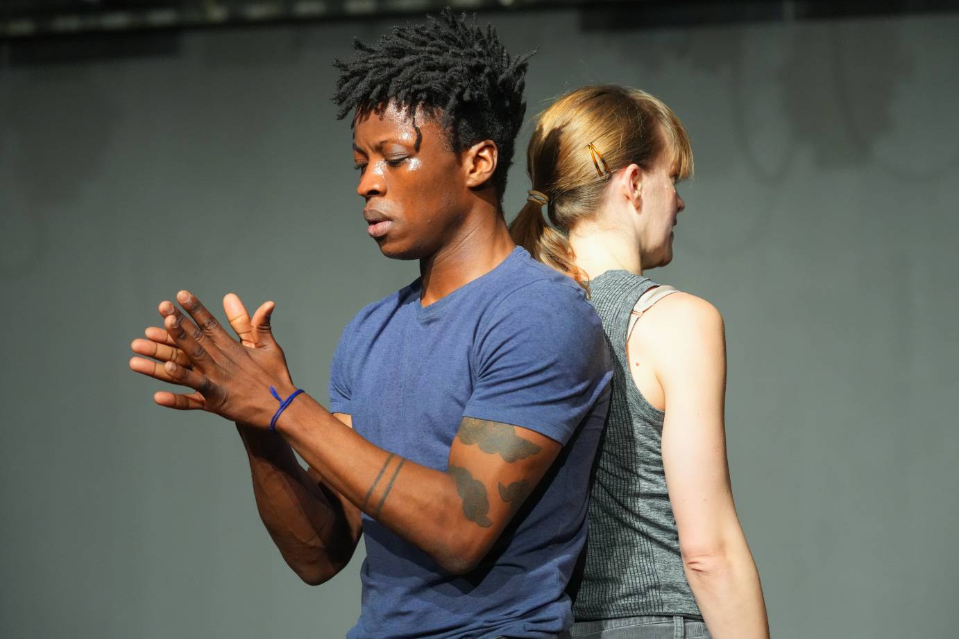 Mufutau Yusuf and Jamie Scott stand back to back. Jamie's face is hidden but she appears to be speaking.Mufutau glances at his hands while rubbing them together. We notice dark tattoos on his arms that look like birds.