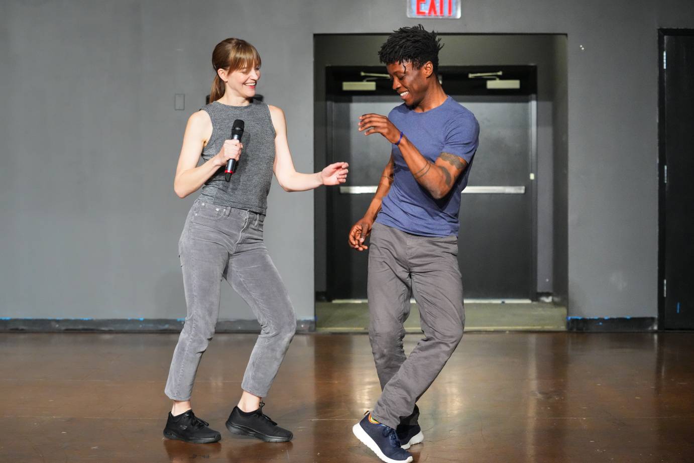 Jamie Scott, a white woman with red hair and Mufutau Yusuf, a Black man with black hair smile and appear to be in the midst of a jitter bug dance. She is dressed in all grey with black sneakers, he in a blue shirt and gray pants with black sneakers. The setting is casual.