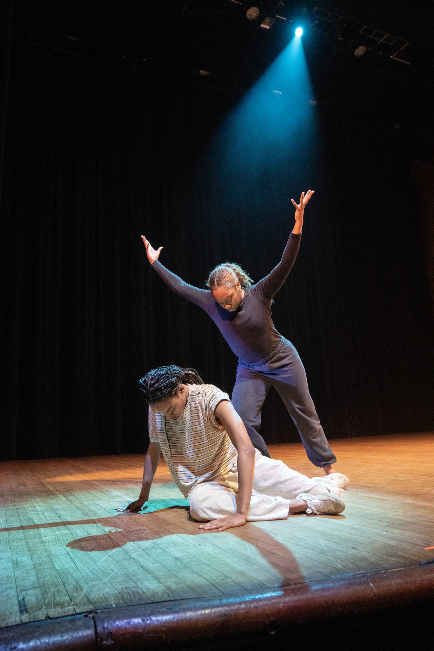 a Black woman with with white pants and a striped shirt and sneakers sits on the floor while another dancer in black stands and leans toward her, arms extended upward