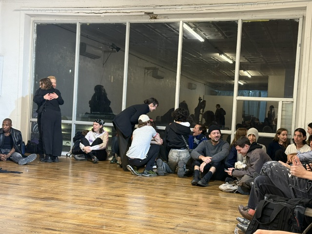 The interior of the PAGEANT space and a view of the darkened picture window reflecting the interior. In front of the window sit audience members; to the left two are standing and hugging.