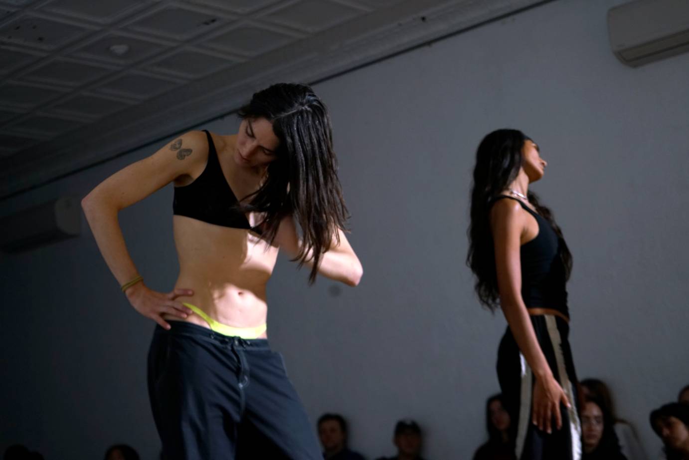 A black and white photo showing a woman lying on her side, her upper body supported by her elbow. Her face is looking away; she is costumed in dark pants, bra, and boots.  