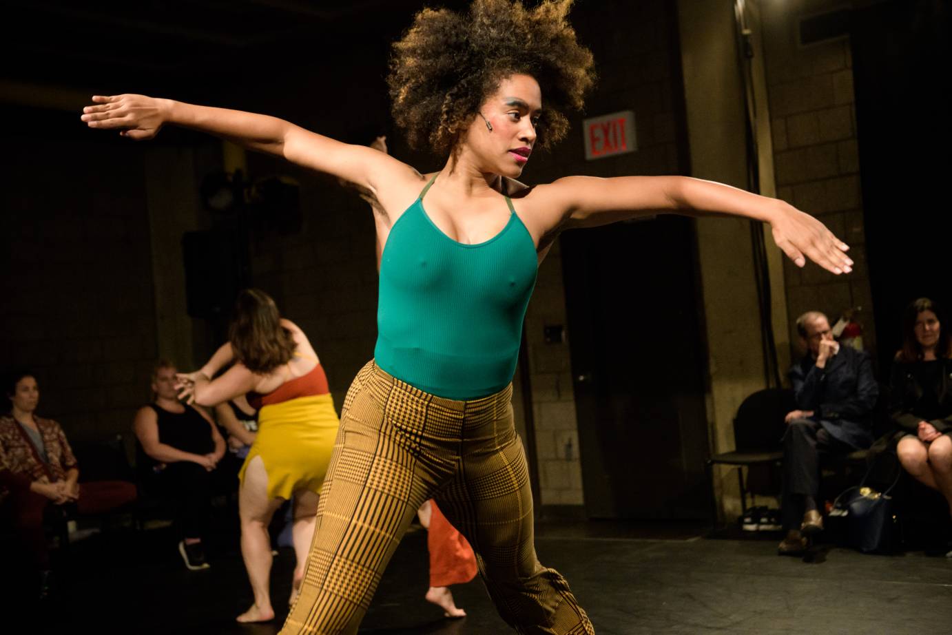 A dancer in a green leotard and plaid brown pants slices her out from her sides.