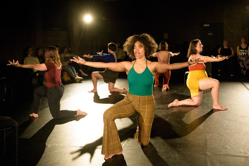 Female-identifying performers speak or sing into headsets. They kneel on one leg facing outward in a circle with their arms outstretched.