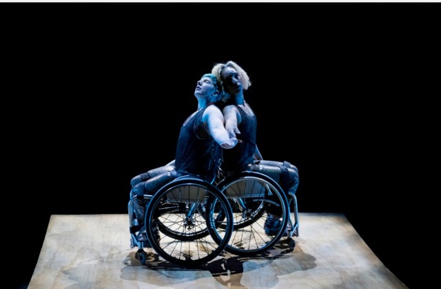 Laurel Lawson and Alice Sheppard are back to back on a wood ramp in their wheelchairs, each with one arm extended toward the camera, heads tilted back. Laurel is a white dancer with cropped blue hair and Alice is a  multiracial Black woman with short curly hair; they both wear shimmery dark sleeveless costumes. Photo by J. Adam Fenster / University of Rochester 