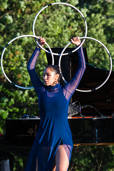 Native American dancer ShanDien LaRance dressed in a short tunic of Royal blue with long sheer sleeves, holds a triangular arrangement of hoops above her head. She is dancing in front of the grand piano outdoors. 