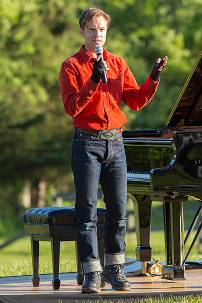 Hunter Noak dressed in a red long sleeved shirt, black fingerless gloves, jeans, black boots, and a belt that appears to be beaded in Native American style addresses the audience, by standing at a microphone beside his grand piano.