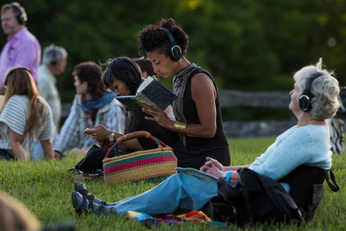 The audience of American Lyric contemplates the music and dance while seated on the grass wearing blue tooth head phones. One of the audience members listens while looking at her notebook. 