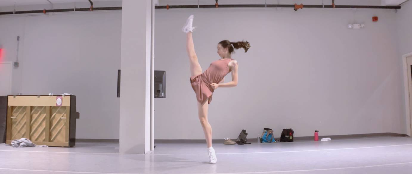 One female dancer in a dusty rose dress kicks her leg forward until it almost touches her nose