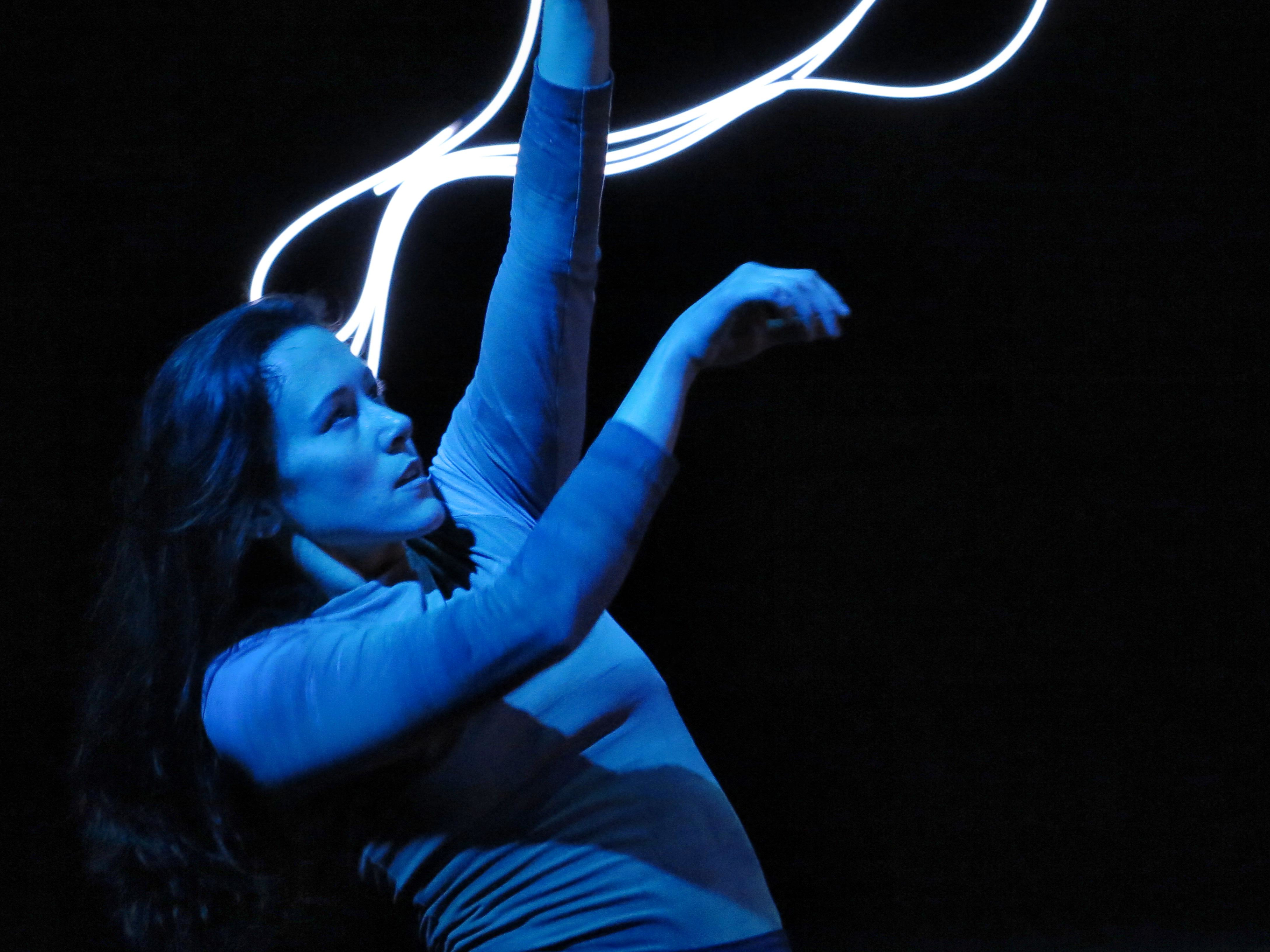 the torso of a female dancer with long brown hair set against the neon light sculpture.. she stands in profile looks towards her outstreched arms