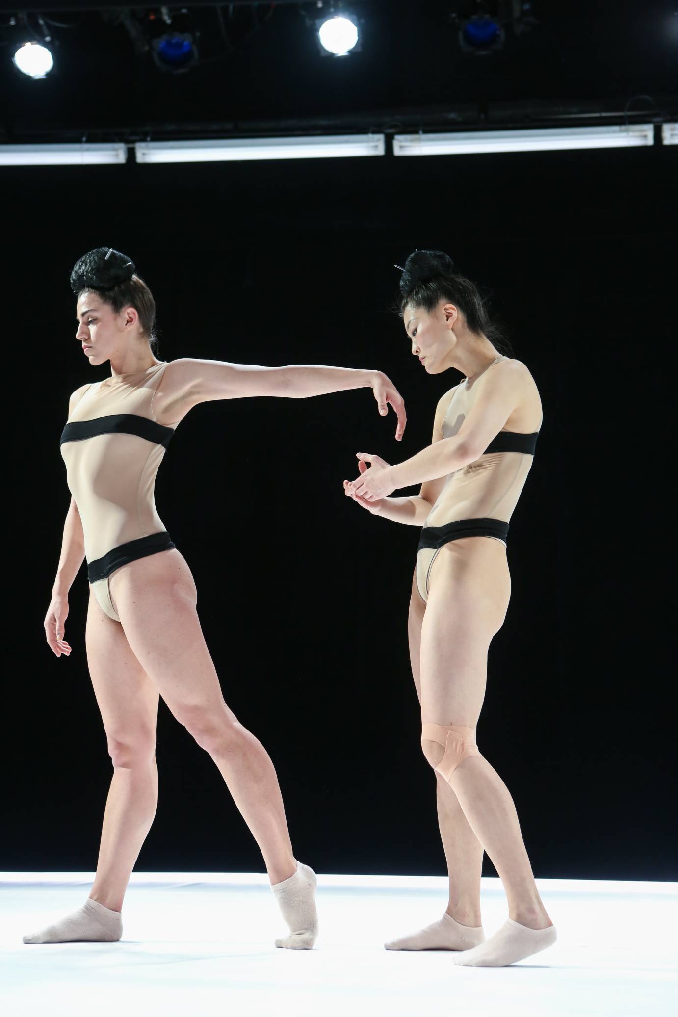 Two women stand in profile; one extends an arm backward while another cups her hands