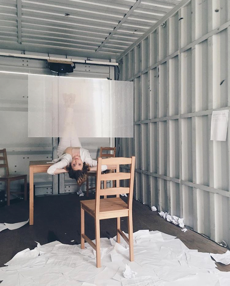 A Woman lies on her back on a table. She has her arms behind her head and looks toward the camera. Lots of white paper is on the floor. A transparent screen hangs above her.