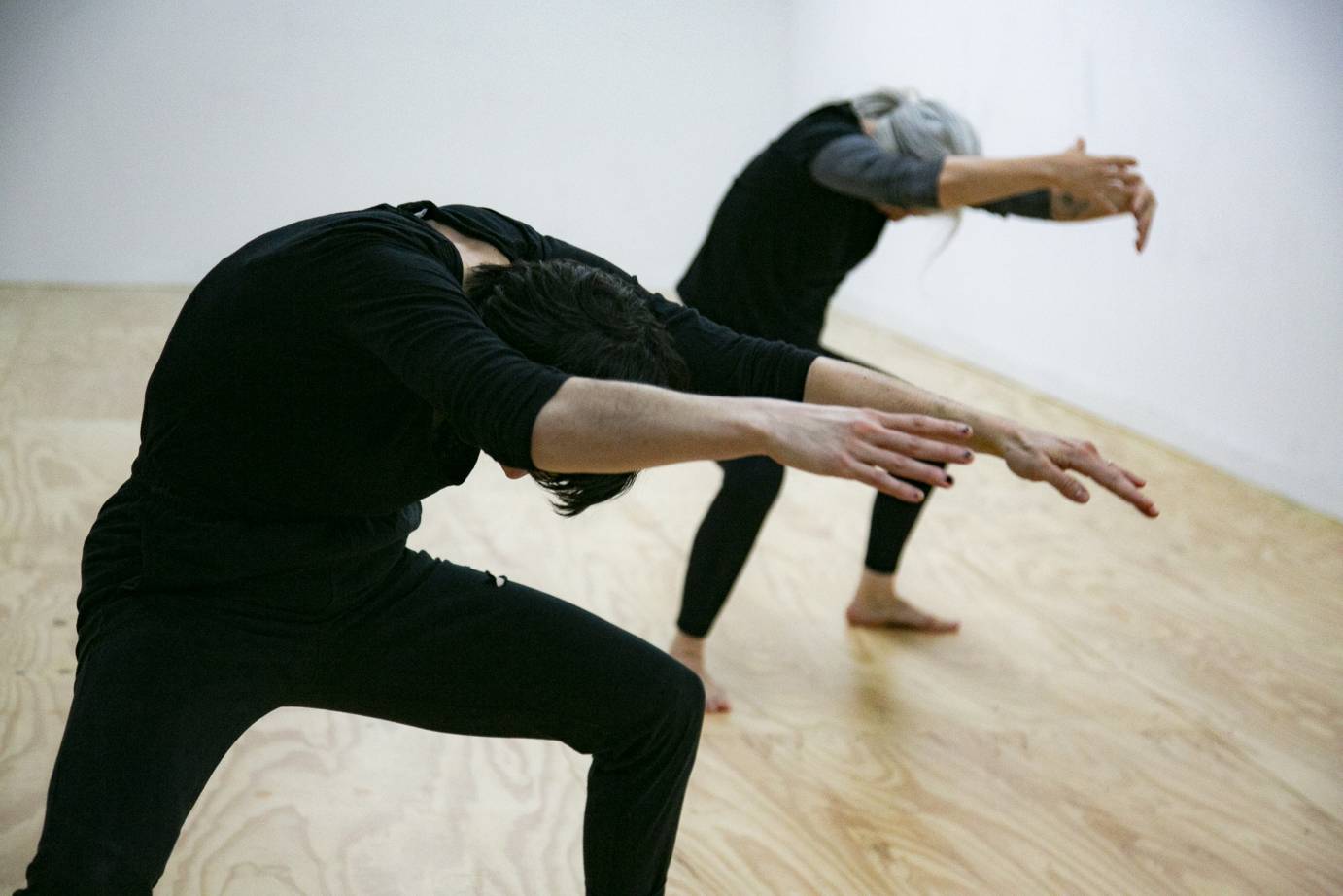 Two dancers facing downstage right diagonal in 2nd position with body curved forward arms outstretched head facing to ground