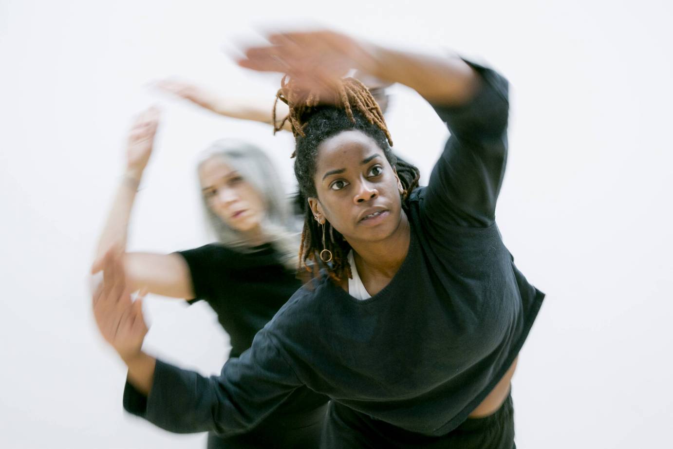 Black woman wearing long sleeved black shirt, faces front. Left arm with bent elbow extends in front of her body. Behind her is a gray haired white women with head slightly tilted to the right but looking forward. Right arm lifted in black short-sleeved shirt.