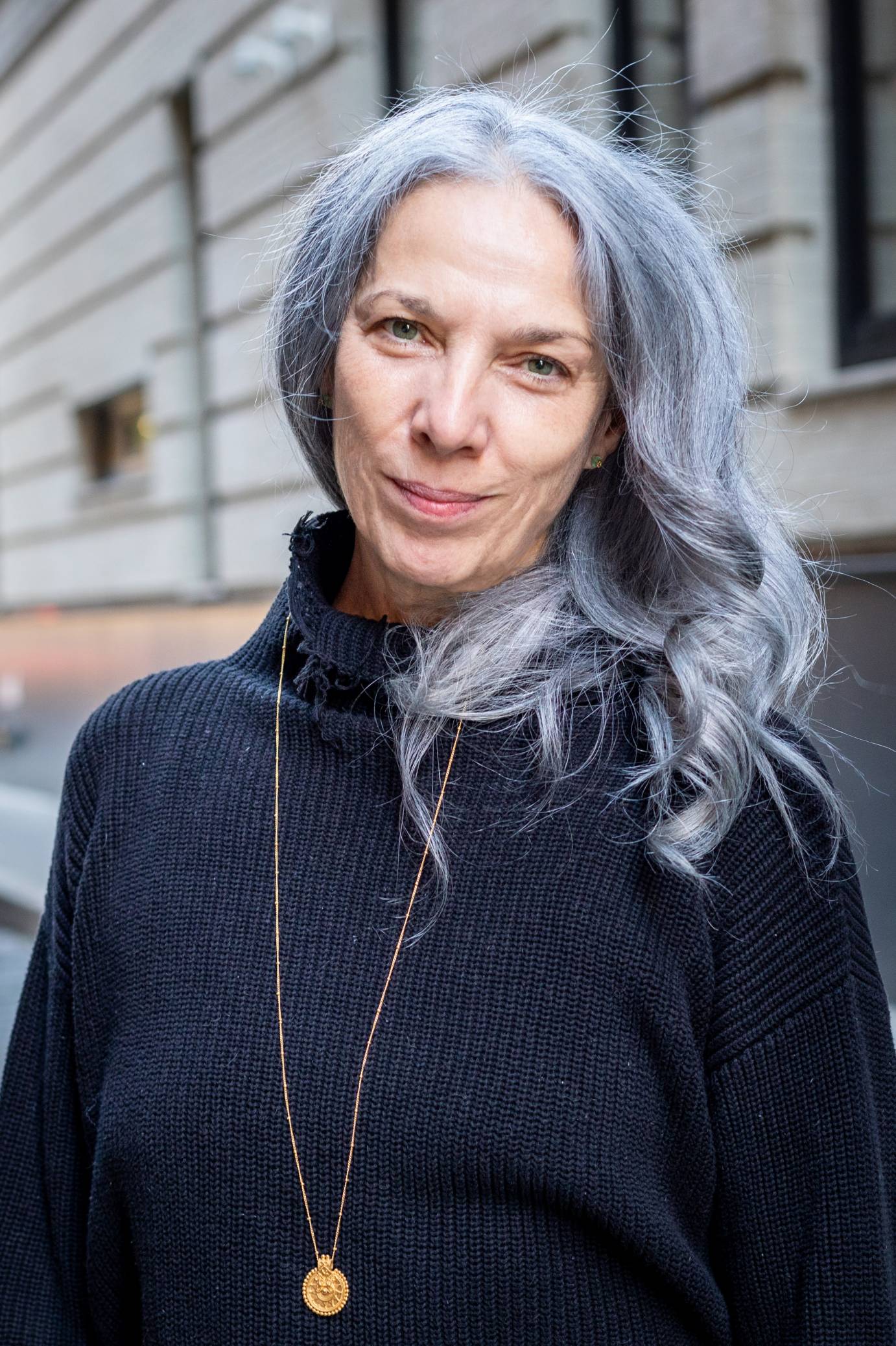 White woman with gray purple hair looking directly into eyes of viewer, a closed mouth smile, waring a purplish turtleneck, a gold medallion hanging from a long gold chain 