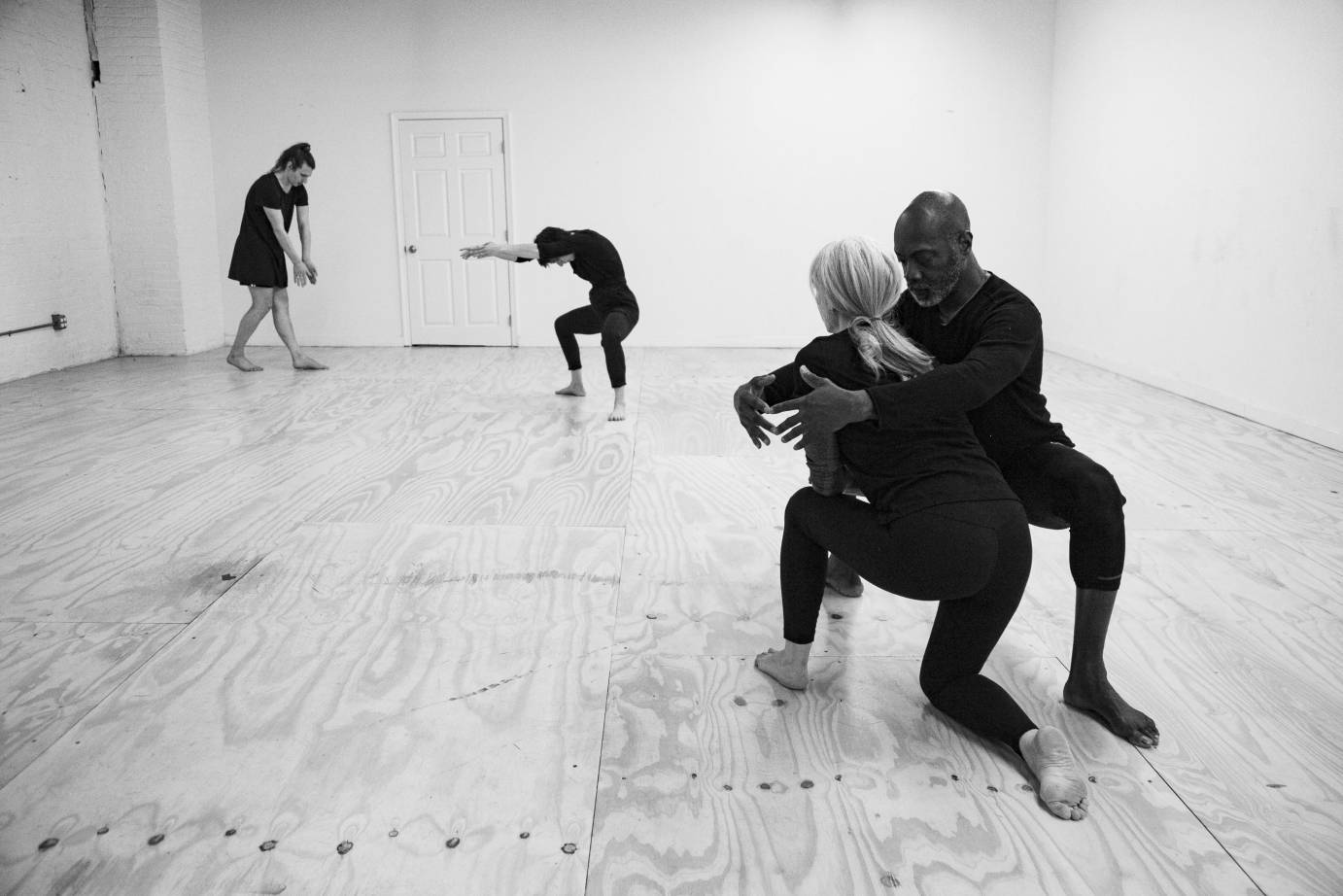 Four dancers: the closest two shows a Black man in a deep 2nd position facing stage right, embracing a gray haired white woman facing upstage. She is looking at two dancers, the farthest left standing upright with head lowered, the other dancer in a deep 2nd position head folded forward and arms in a soft forward 2nd.