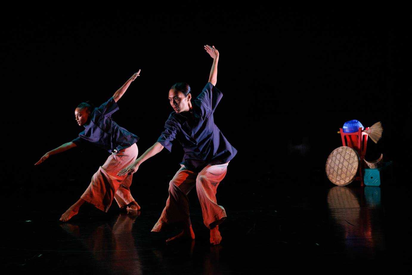 Two women in unison dressed in blue shirts and loose reddish pants extend their arms on the diagonal while looking over their lower arms.
