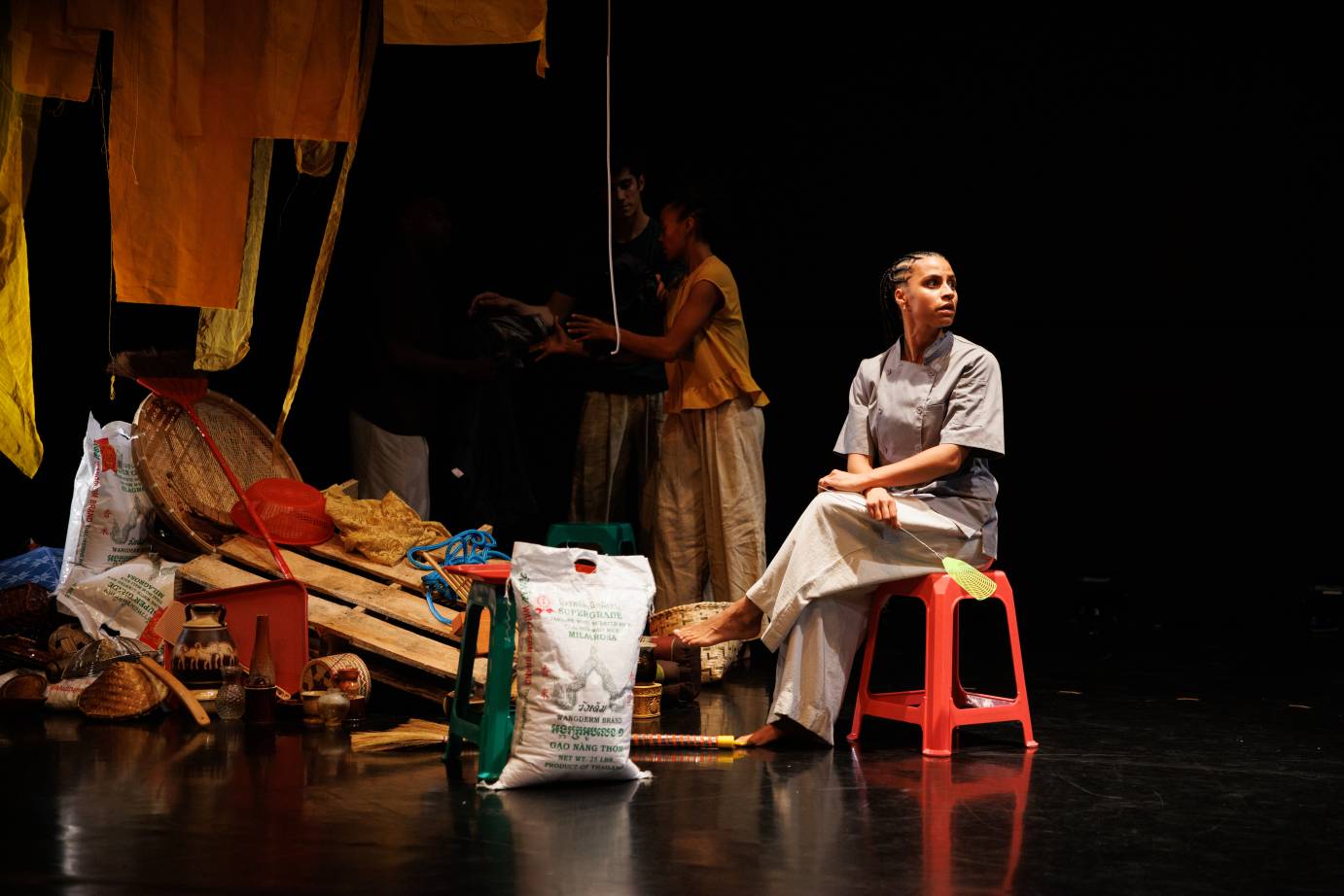 A woman with a head of braided hair sits cross-legged on a red stool with flyswatter in hand. In front of her spills items from a commercial kitchen including a huge white bag of rice.