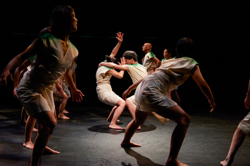 A group of dancers in white robe-like costumes. Two dancers face one another and bend their legs deeply. Their hands block their faces.