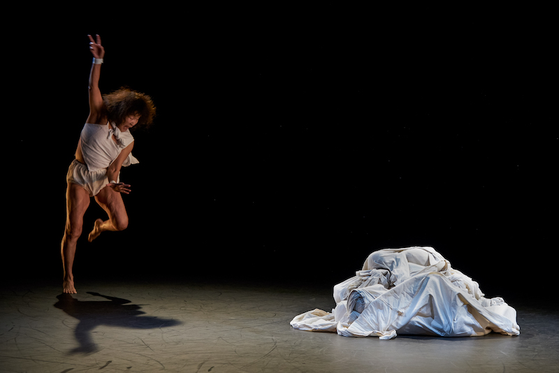 Kei Takei in loose briefs and tank top opens her mouth wide and is in mid-movement. She stands on one leg and one arm is outstretched above her. A heap of white fabric lays to her left.