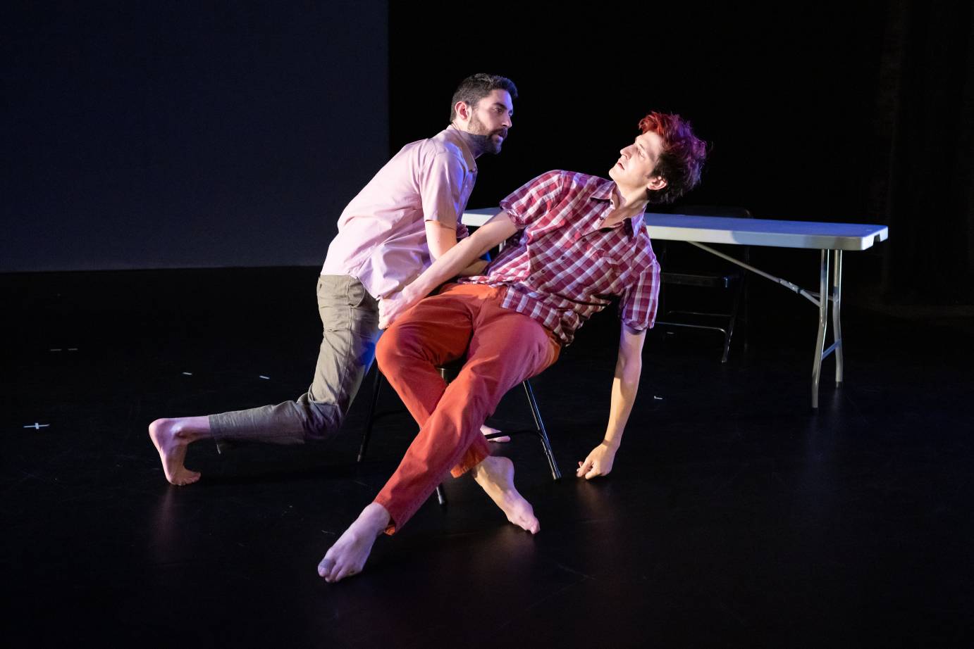 Two men stare into each other's faces as they lean back in a chair close to a collapsible. One man has red dye in his hair and is dressed in a red plaid shirt and red jeans. The other man, who is bearded, is dressed in a polo shirt and everyday pants. table. 