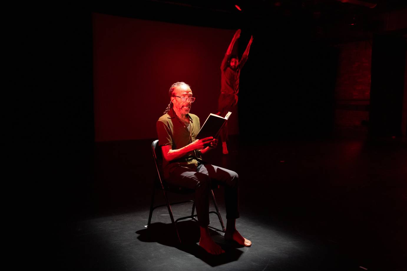 A long-haired man wearing red-framed glasses sits in a collapsible chair reading from a dark bound book. A bearded man is stretched long upstage.