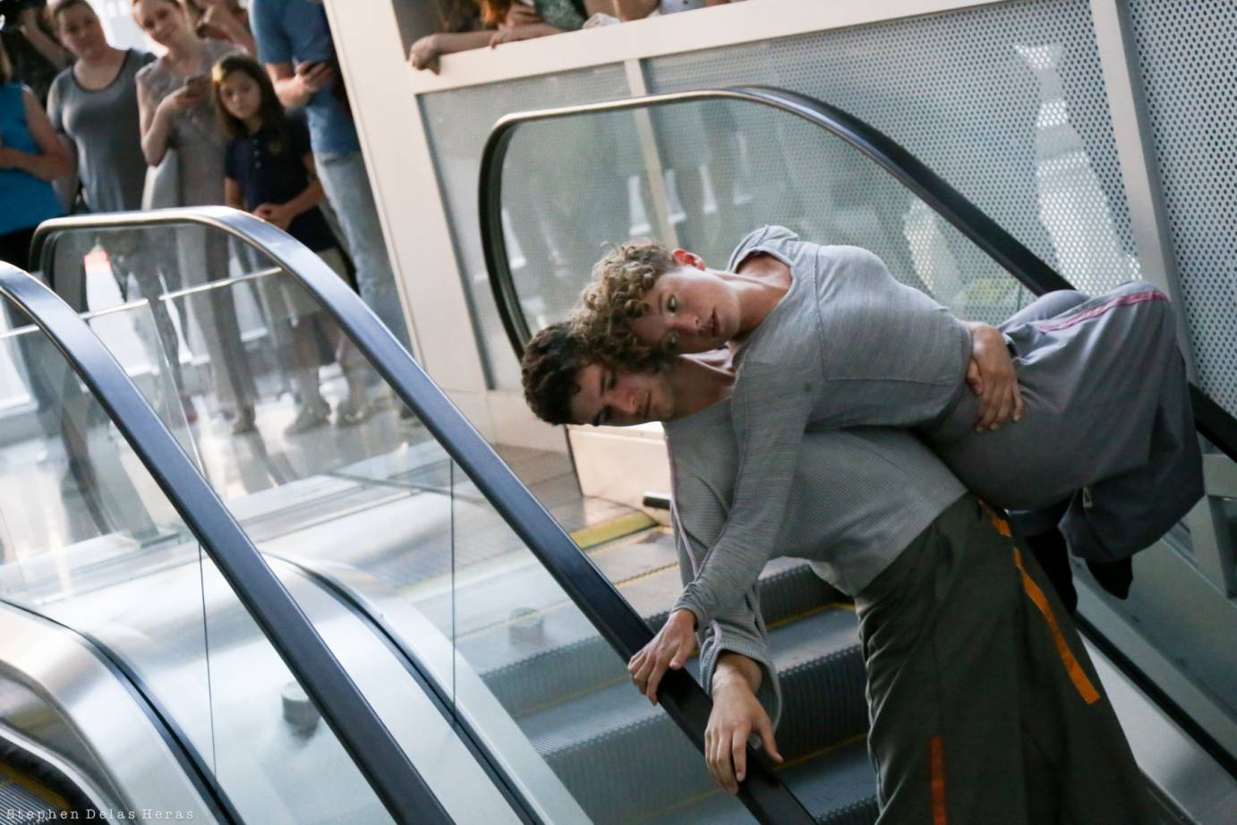 Two dancers in grey dance on a escalator. One performer picks up the other.