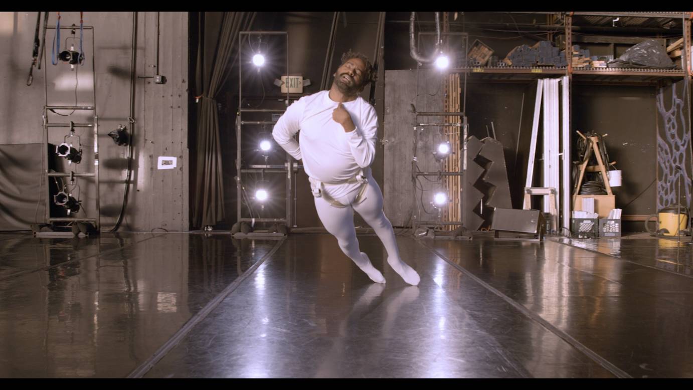 Balancing on only the tops of his toes, Jerron leans his whole body forward, and gives his weight into the cables which extend from above to his waist. He is a dark-skinned Black man with a beard and twisted black hair, wearing all white in a black studio space. His right arm bends behind his back as his left clings to his chest. Eyes closed and face relaxed, Jerron is in a moment of pensive release. Stage lights and miscellaneous equipment scatter across in the background.