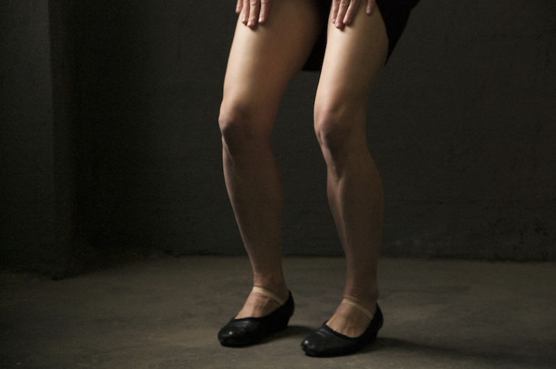A close of two bare legs bent at the knees with their feet in black flats with straps