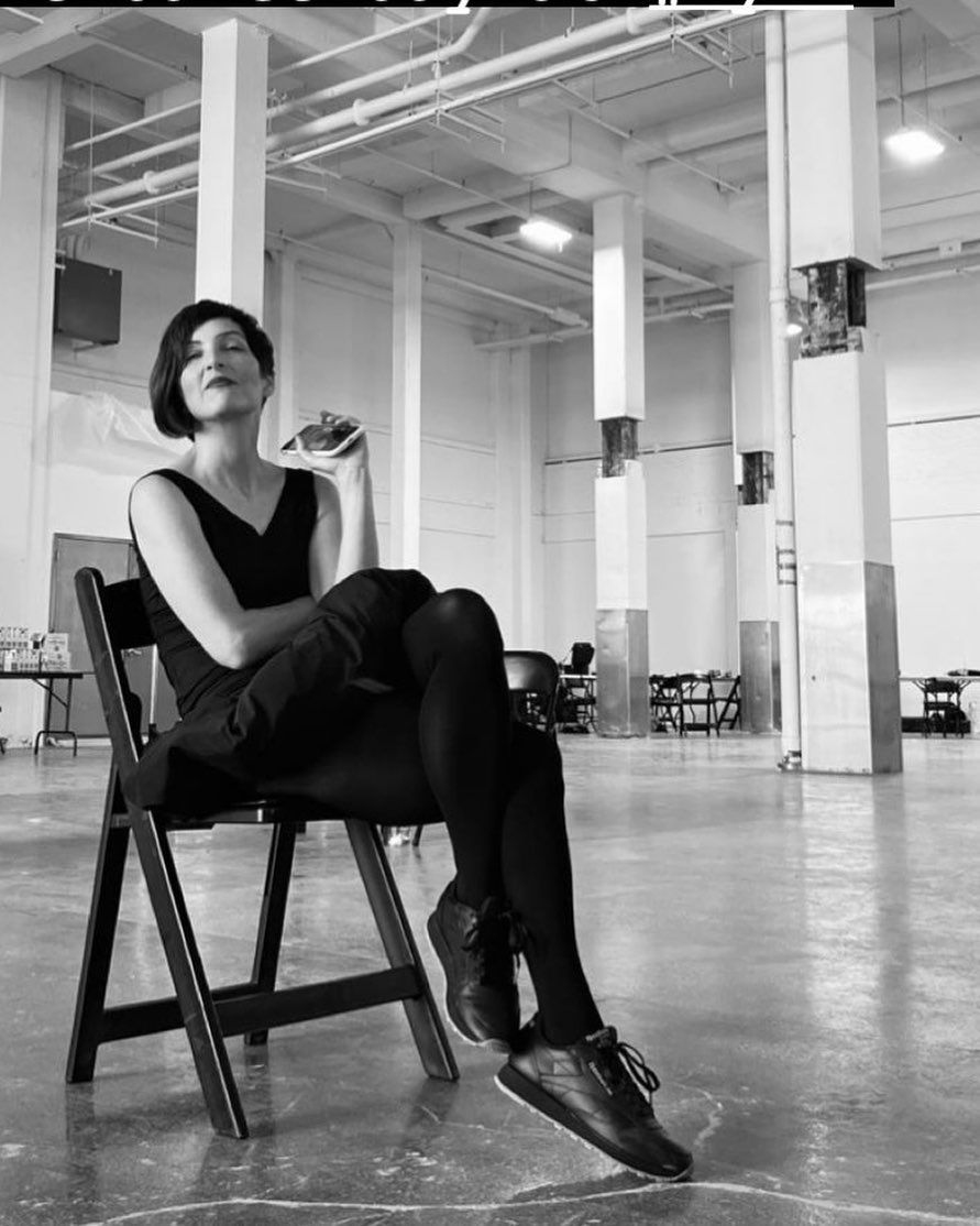 Laura Peterson sitting on a chair in a studio, black and white photo, she smiles with her cell phone in one hand other arm on her lap legs crossed, chin lifted, she sports an assymetrical bob
