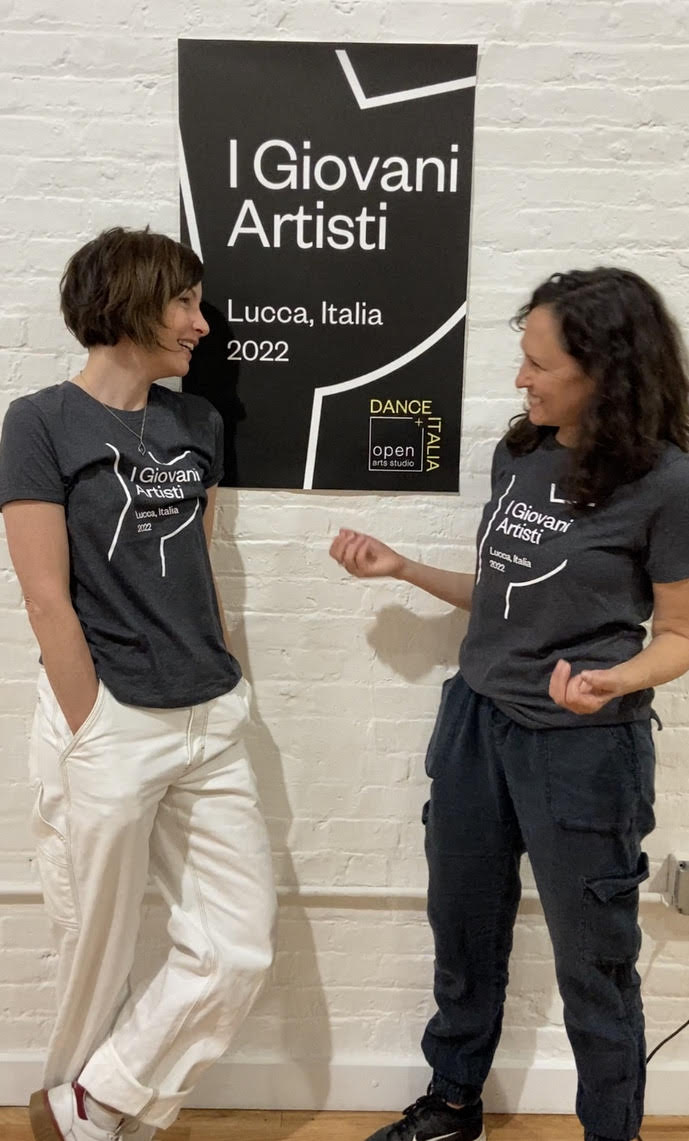 laura peterson and stefanie nelson in t shirts and relaxed pants laugh in front of the poster  for the young artists Lucca, Italia