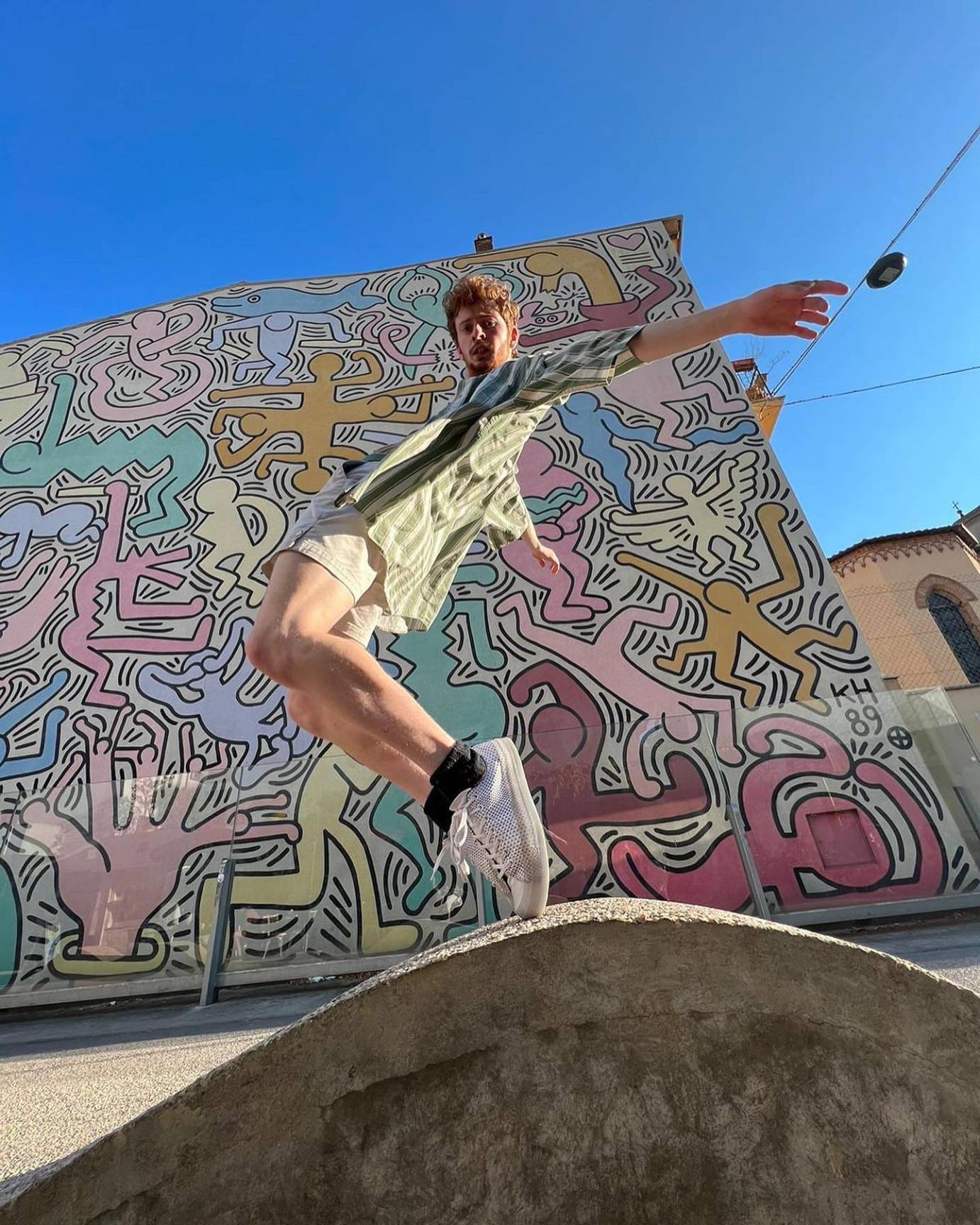 young teen fair skinned male dancer with curly light hair and a beard and mustache. He wears light shorts and a green and white striped top as he dances in front of the Keith Haring mural