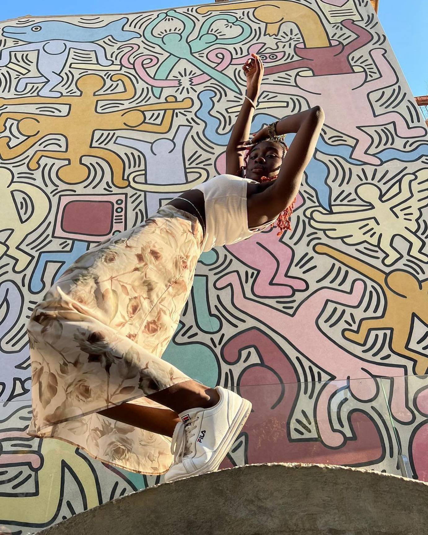 a young black teen girl in light clothing and sneakers poses in front of a Keith Haring mural which radiates dance energy