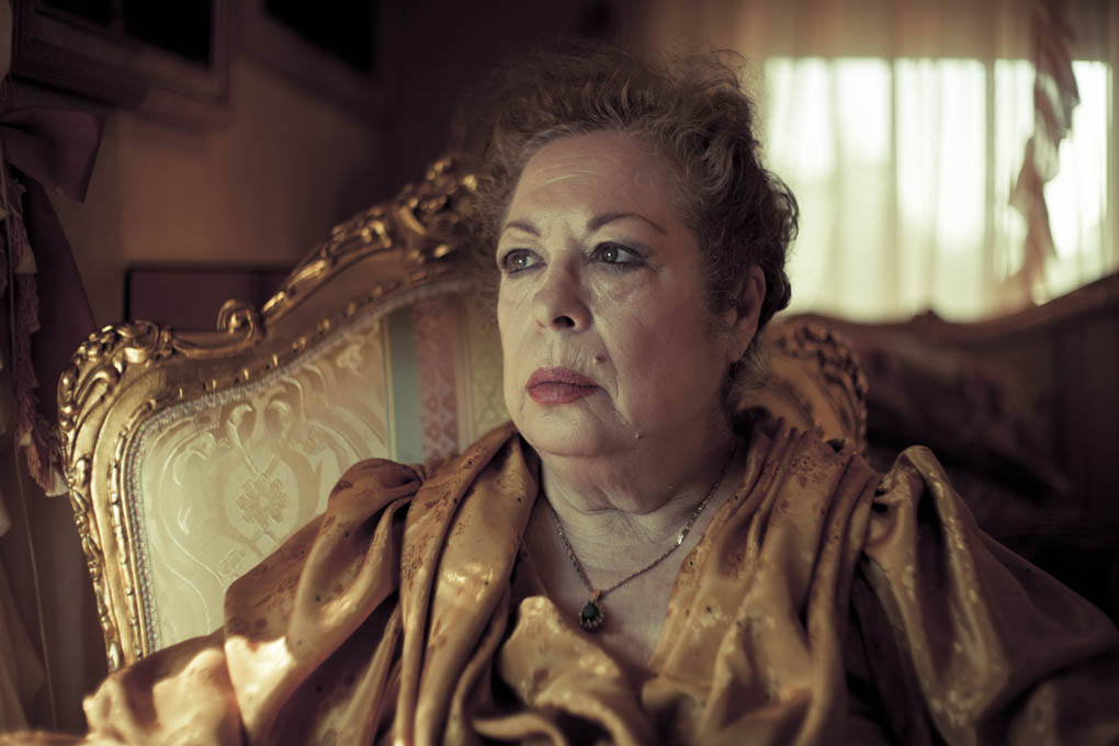 A close of La Chana who sits in an ornate upholstered chair. She stares away from the camera.