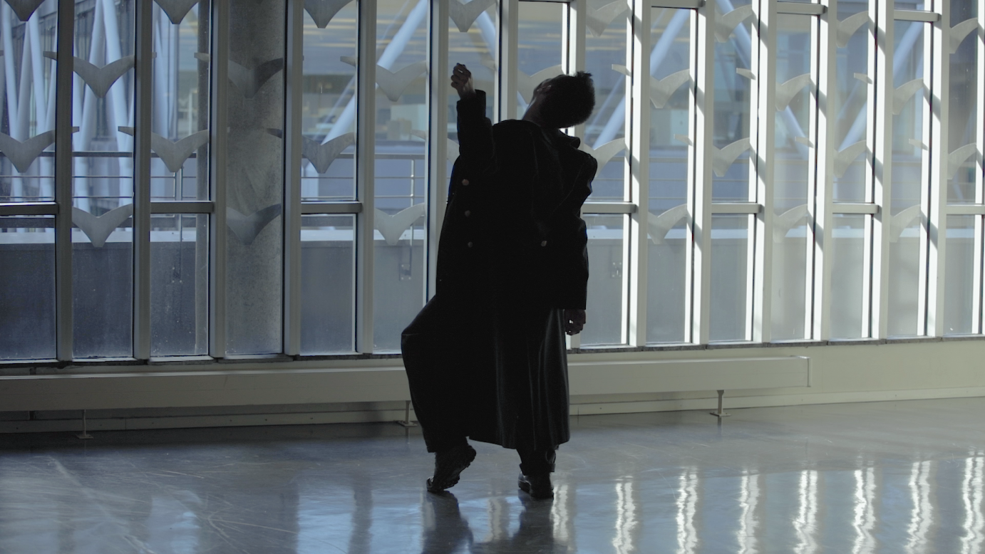 Jens Jeffrey Trinidad, in a long jacket, extends his arm with a cupped hand while in an airport.