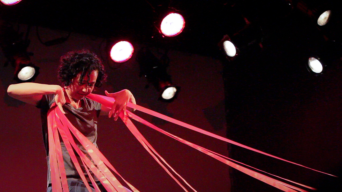 Pele Bauch a Chinese Hawaiian woman with curly black hair in a gray tshirt and pants dances with long red tape around her neck 